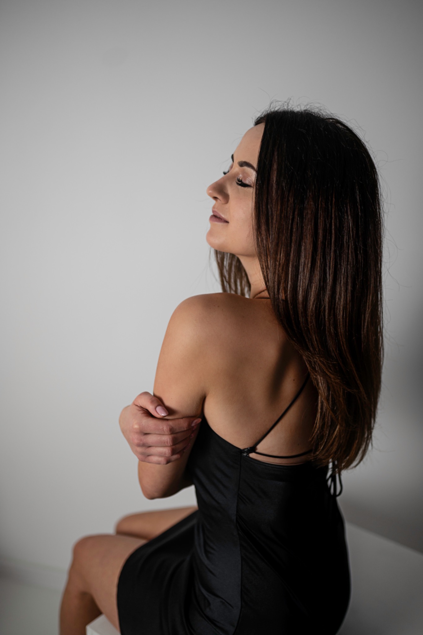 Kobieta w czarnej sukience z cienkimi ramiączkami, siedząca bokiem, z zamkniętymi oczami i rękami skrzyżowanymi na ramieniu, na jasnym tle.