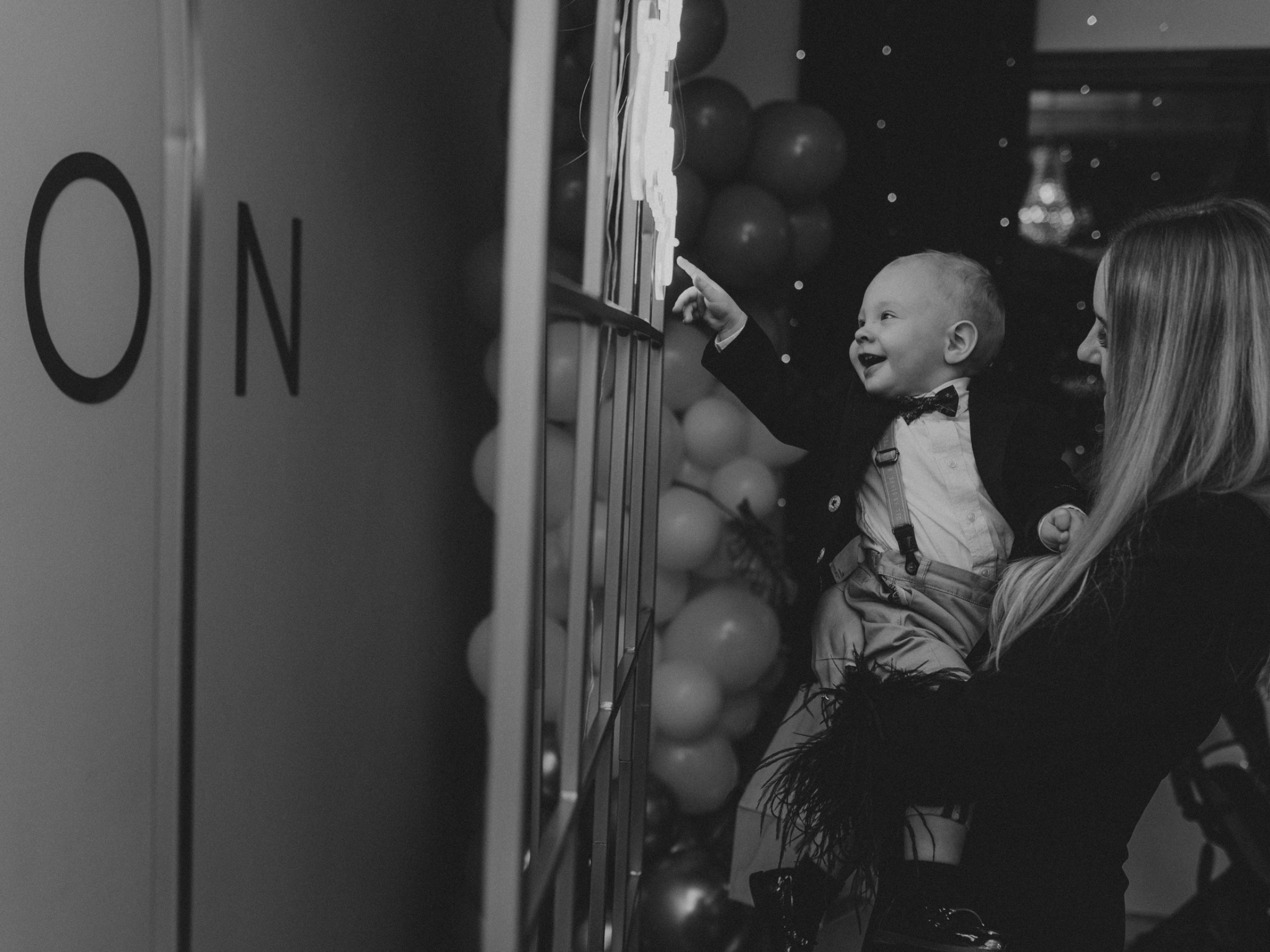 Uśmiechnięty chłopiec w eleganckim stroju, trzymany na rękach przez kobietę, wyciąga palec w stronę dekoracji świetlnej na tle balonów podczas uroczystości.