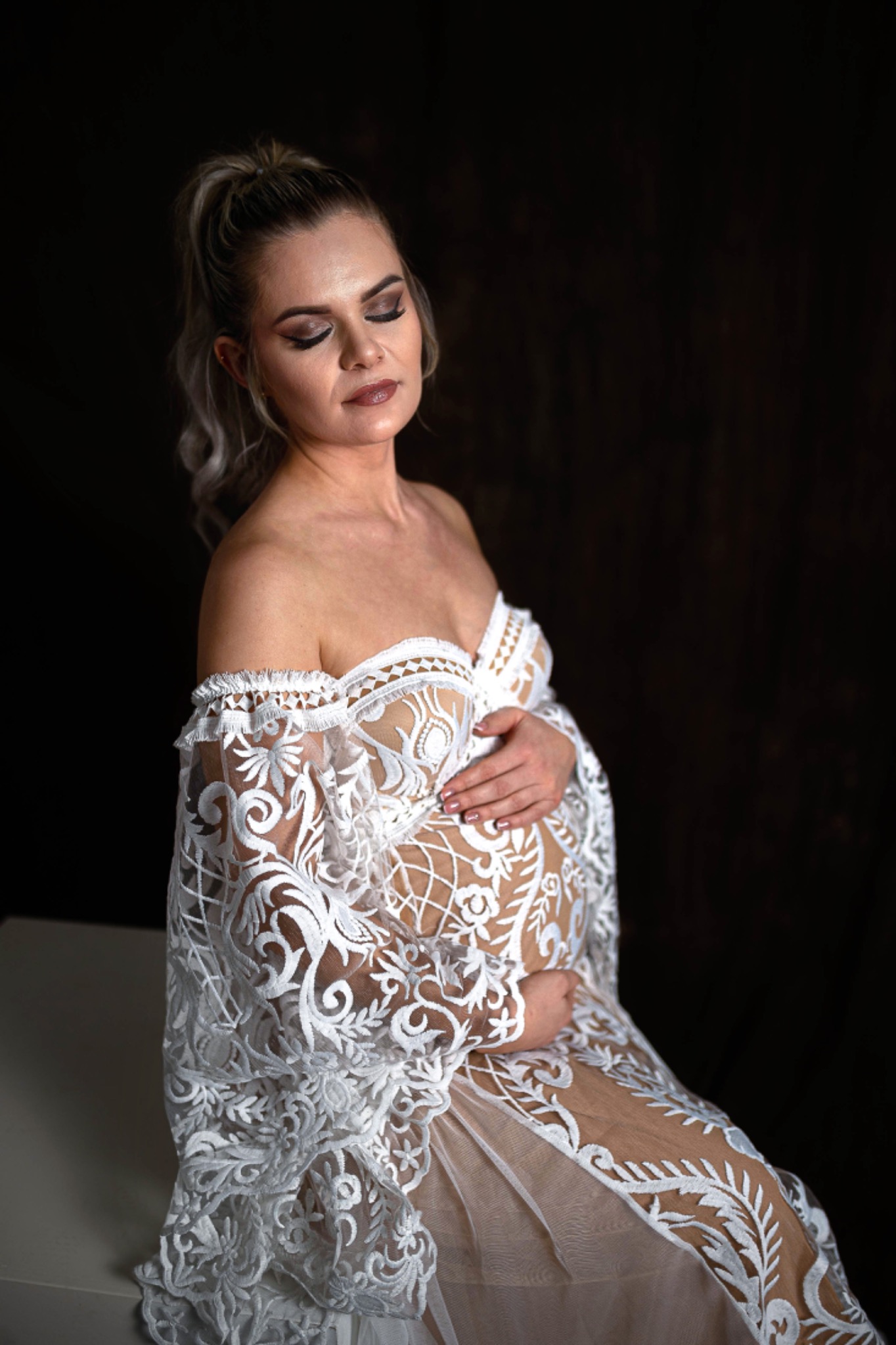 Kobieta w ciąży, w białej, koronkowej sukni, z rękami na brzuchu, pozuje na ciemnym tle.