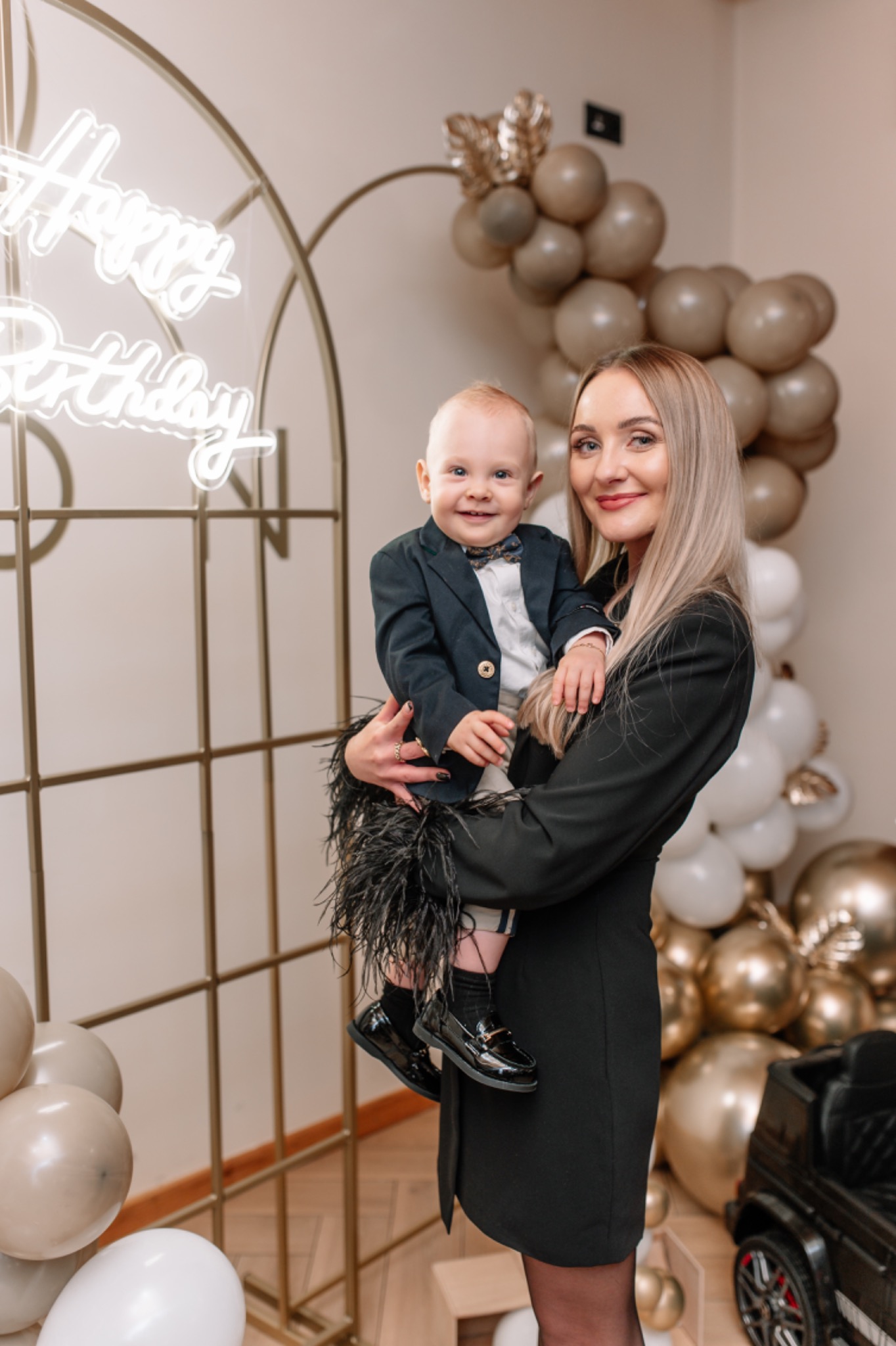 Uśmiechnięta mama trzymająca na rękach elegancko ubranego synka na tle dekoracji urodzinowych z balonów i neonowym napisem 'Happy Birthday'.