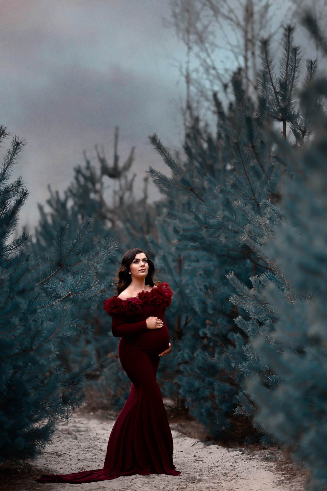 Kobieta w bordowej sukni ciążowej z falbanami pozuje na tle zimozielonych drzew, spoglądając w górę.