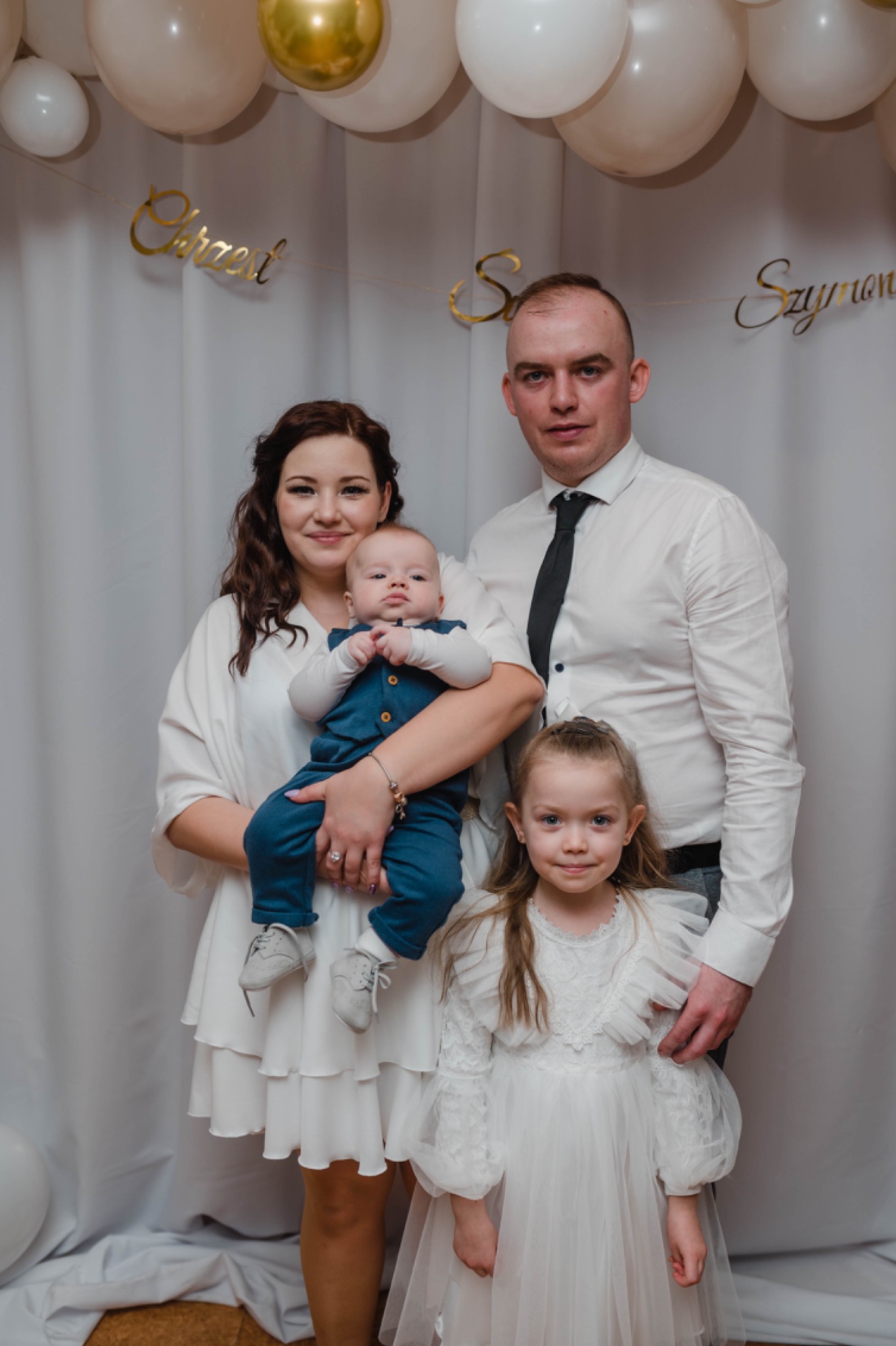 Uśmiechnięta rodzina: mama trzyma niemowlę w niebieskim ubranku, obok stoi tata w koszuli i krawacie oraz mała dziewczynka w białej sukience, w tle białe balony i złoty napis 'Chrzest Szymona'.