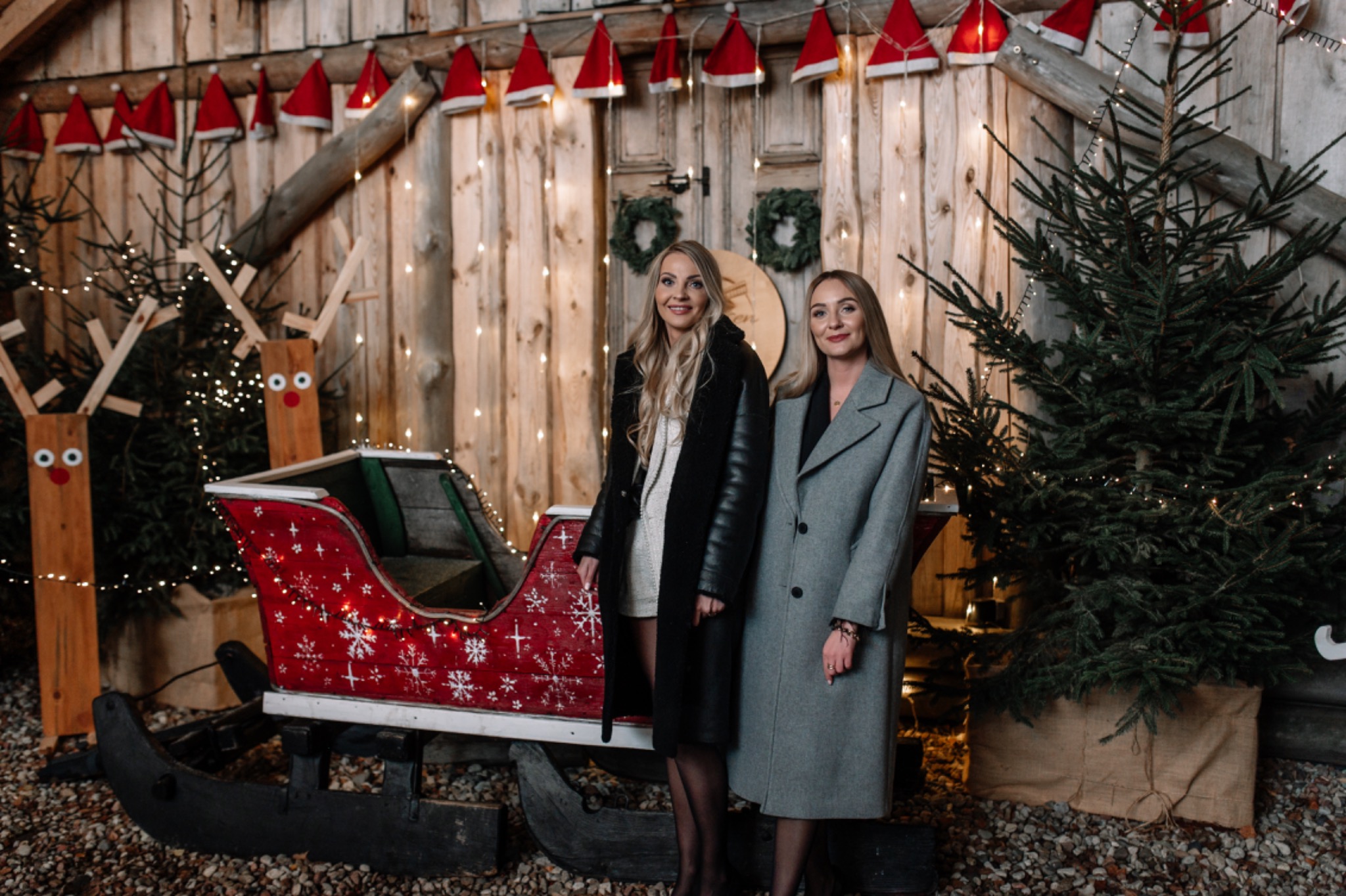 Dwie uśmiechnięte kobiety w płaszczach pozują obok czerwonych sań z motywem śnieżynek, drewnianych reniferów i choinki, na tle drewnianej ściany ozdobionej lampkami i czapkami Mikołaja.
