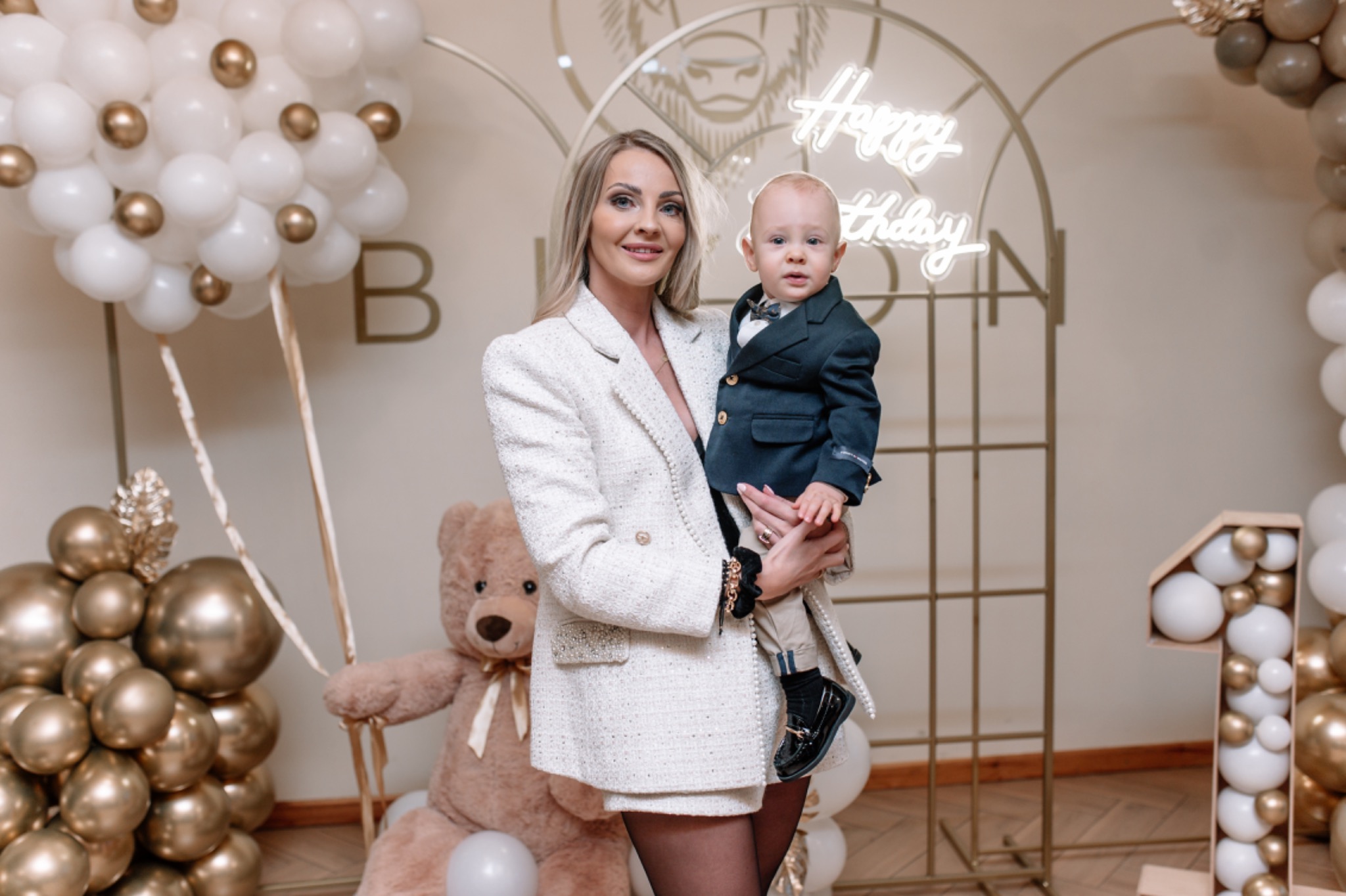 Elegancka kobieta w białym tweedowym kostiumie trzyma na rękach ubranego w granatowy garnitur chłopca na tle dekoracji urodzinowych z balonów, pluszowego misia i neonowego napisu 'Happy Birthday'.