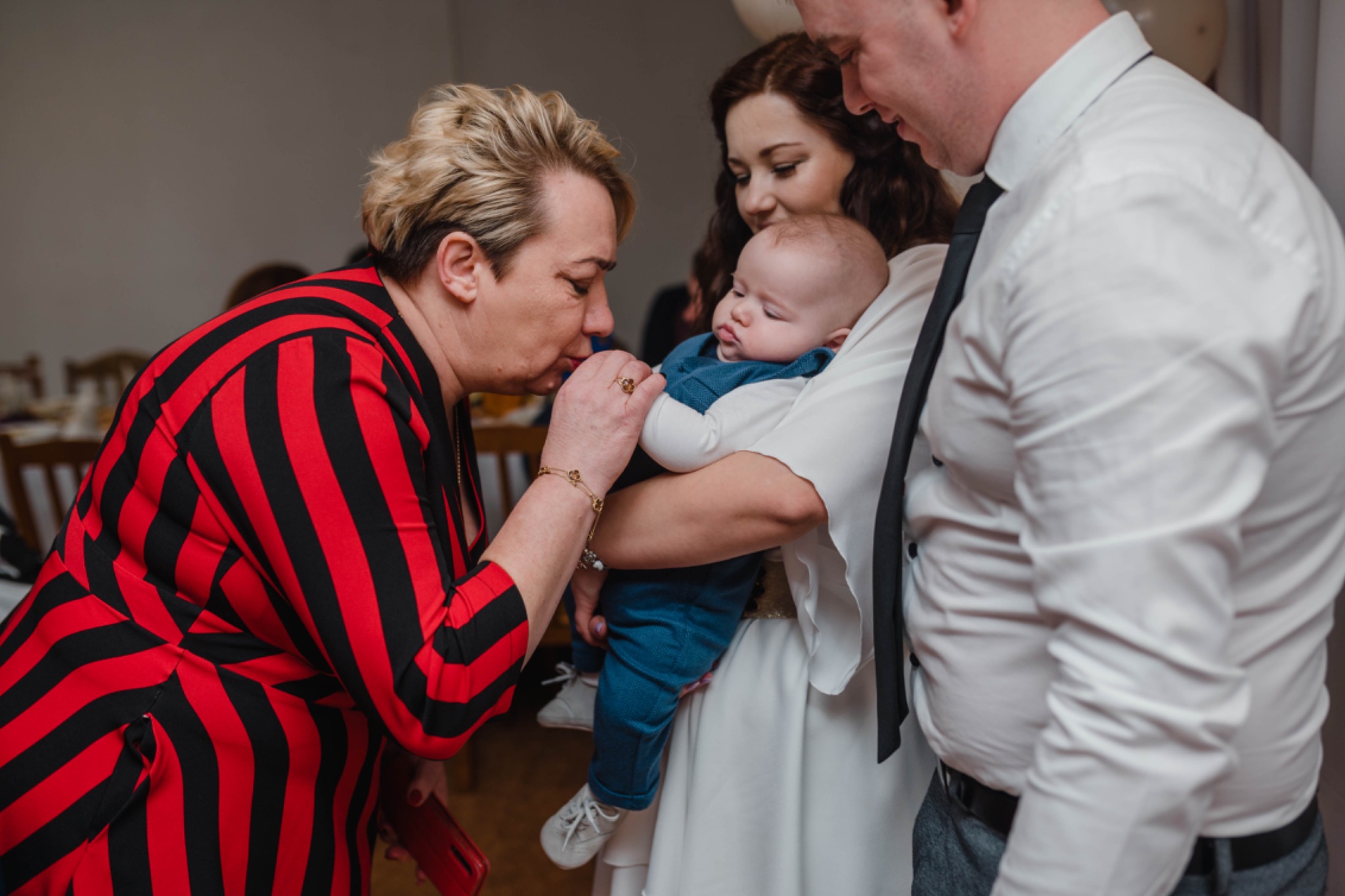 Kobieta w czerwono-czarnej sukience w paski całuje dłoń niemowlęcia trzymanego przez rodziców podczas uroczystości.