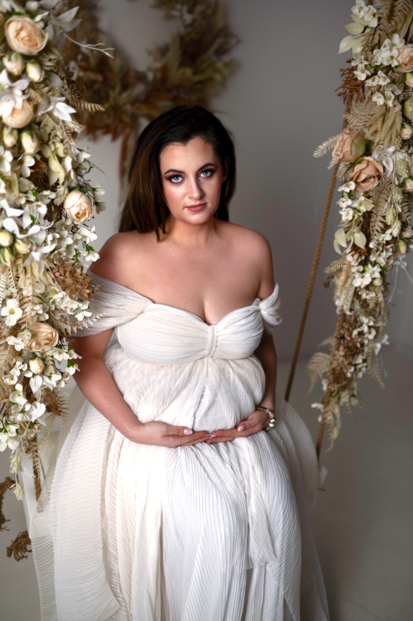 Kobieta w ciąży w białej sukni siedzi na huśtawce udekorowanej kwiatami, delikatnie trzymając dłonie na brzuchu, z uwagą skierowaną w obiektyw.