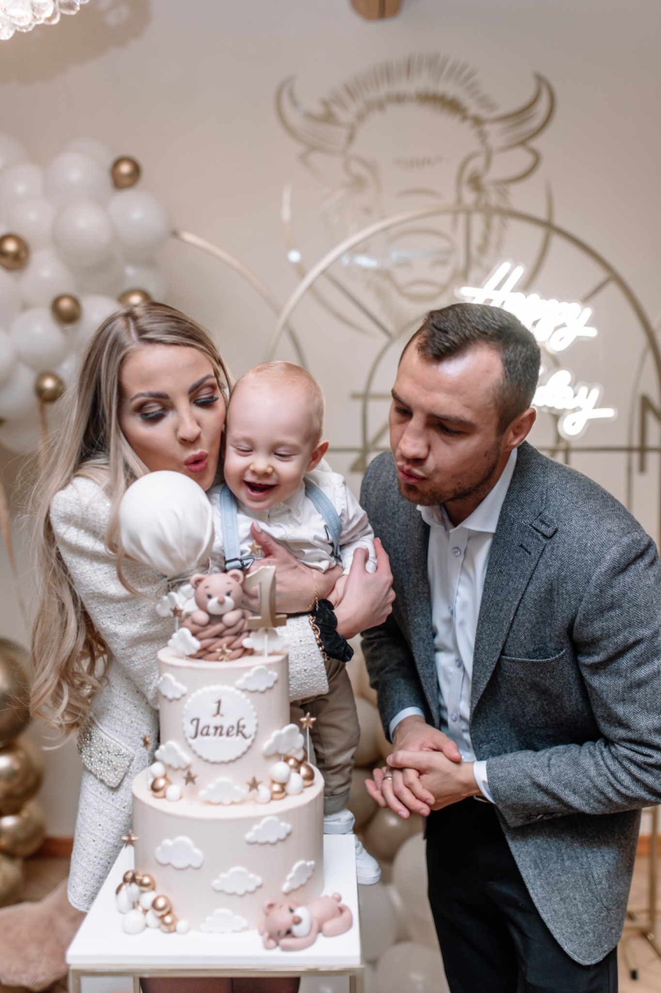 Rodzina świętuje pierwsze urodziny dziecka z tortem ozdobionym chmurkami i misiem, na tle balonów i neonowego napisu.