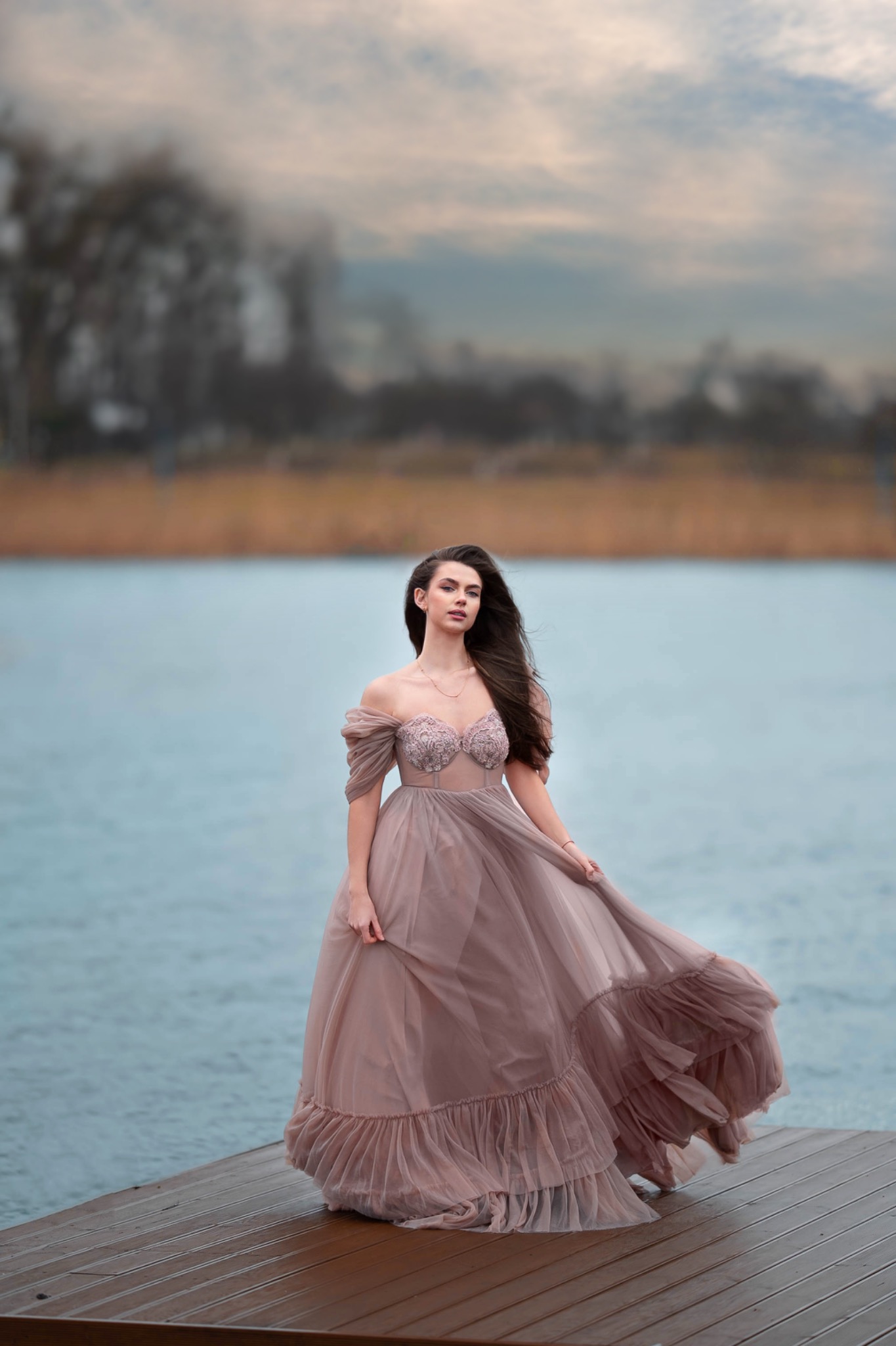 Kobieta w zwiewnej, pudrowo-różowej sukni stoi na drewnianym pomoście nad wodą, z rozpuszczonymi, ciemnymi włosami i delikatnym makijażem, pozując na tle zamglonego krajobrazu.