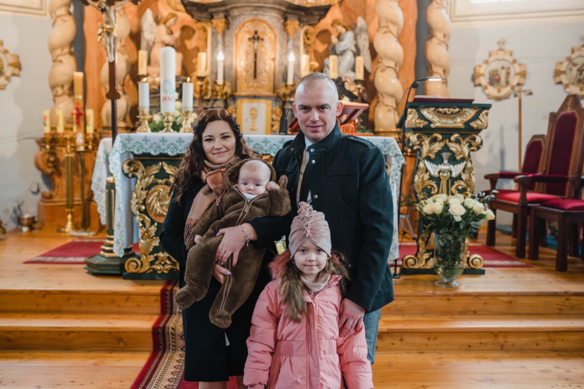 Rodzina pozuje do zdjęcia w kościele na tle ołtarza: rodzice, mała dziewczynka w różowej kurtce i niemowlę w brązowym kombinezonie.