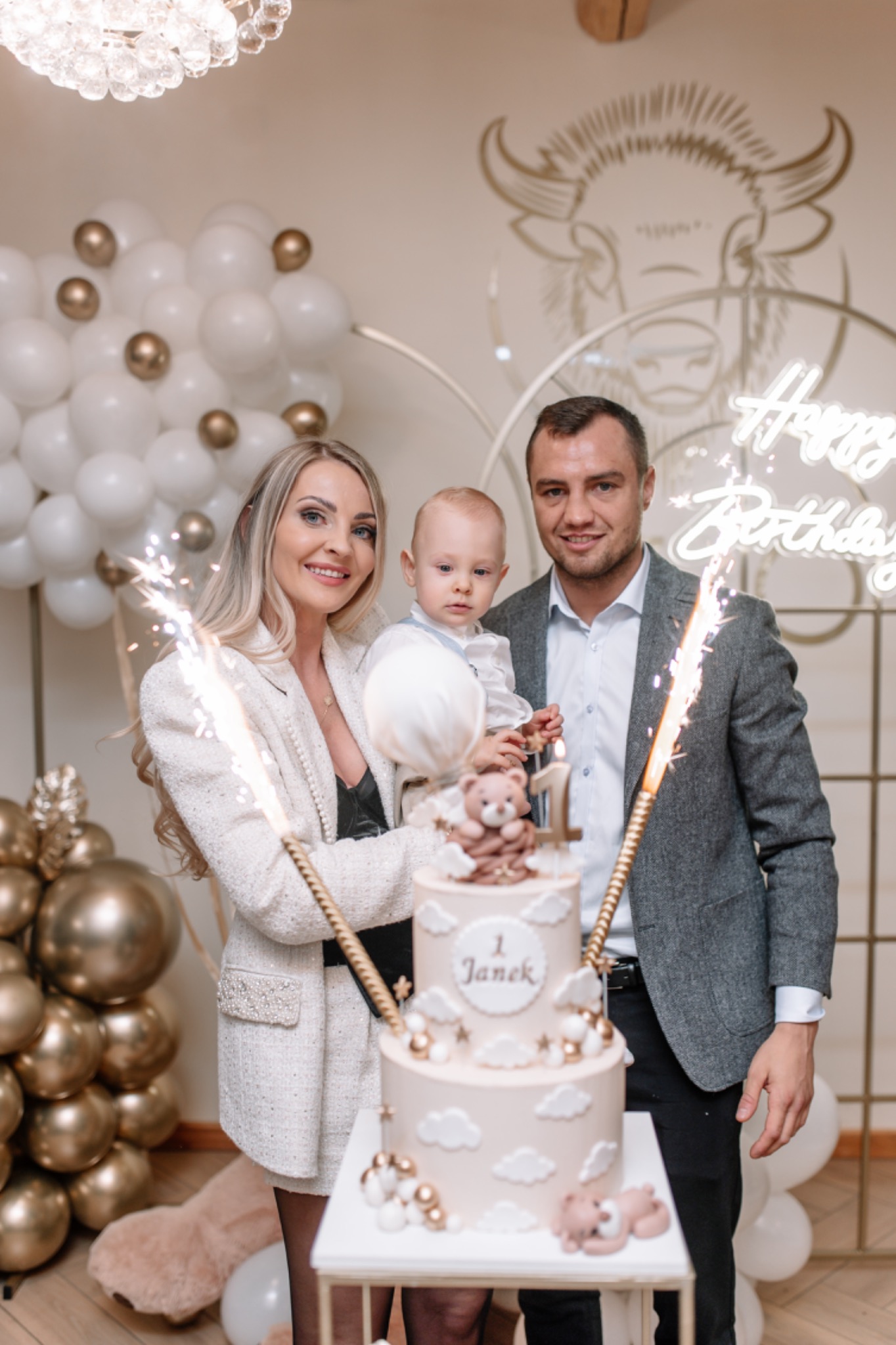 Rodzinne zdjęcie z tortem urodzinowym dla rocznego dziecka, dekoracje balonowe i zimne ognie tworzą świąteczny nastrój.