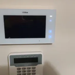 Biały panel wideodomofonu Vidos z podświetlanymi przyciskami i ekranem oraz klawiatura alarmowa Safes instalowane na beżowej ścianie wewnątrz budynku, widoczny fragment ciemnobrązowej framugi drzwi...