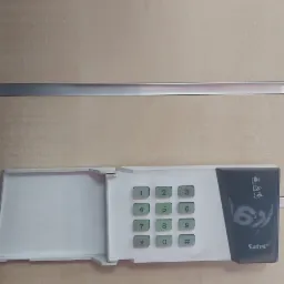 Klawiatura alarmowa Satel z otwartą klapką ochronną, zamontowana na drewnianej ścianie z metalowymi listwami.