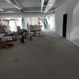 Wnętrze w trakcie wykańczania: widoczne surowe ściany, instalacje pod sufitem, materiały budowlane i narzędzia, w tle postać pracownika.