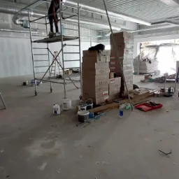 Instalator na rusztowaniu montuje instalację pod sufitem w hali, widoczne skrzynki i narzędzia.