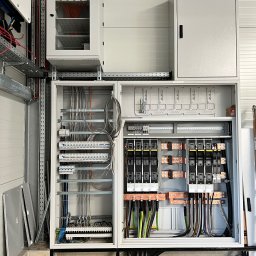 F.H.U LED ELEKTRO - Otwarta, duża szafa rozdzielcza z widocznymi bezpiecznikami, przewodami i listwami zaciskowymi, dodatkowa szafka z przeszklonymi drzwiami powyżej, na tle białej ściany.