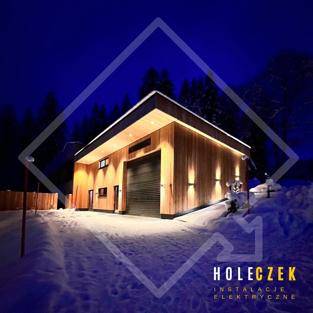 Nowoczesny budynek z drewnianą elewacją oświetlony nocą, na śniegu, z widocznym logo firmy 'HOLECZEK INSTALACJE ELEKTRYCZNE' w dolnej części zdjęcia.