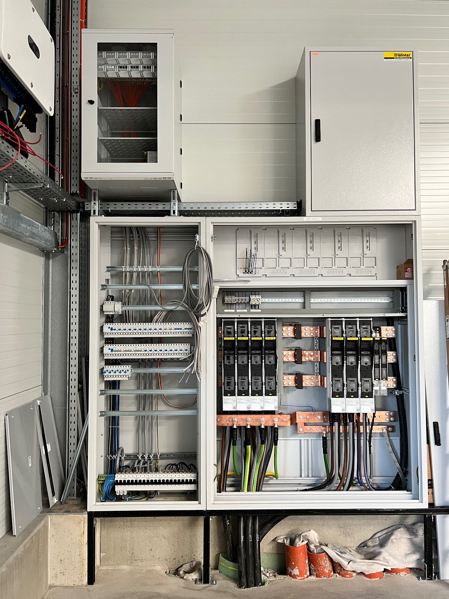 Otwarta, duża szafa rozdzielcza z widocznymi bezpiecznikami, przewodami i listwami zaciskowymi, dodatkowa szafka z przeszklonymi drzwiami powyżej, na tle białej ściany.