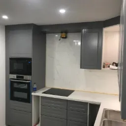 Wnętrze kuchni w trakcie montażu: szare szafki kuchenne z wbudowanym piekarnikiem i płytą grzewczą, marmurowe płytki na ścianie, widoczne elementy montażowe i butelka wody mineralnej.