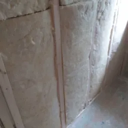 Wnętrze w trakcie ocieplania: wełna mineralna między drewnianymi belkami, surowa podłoga w tle.