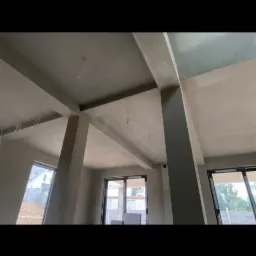 Surowe wnętrze budynku w trakcie prac wykończeniowych: widoczne szare ściany, betonowe filary, okna oraz wystające przewody elektryczne z sufitu.