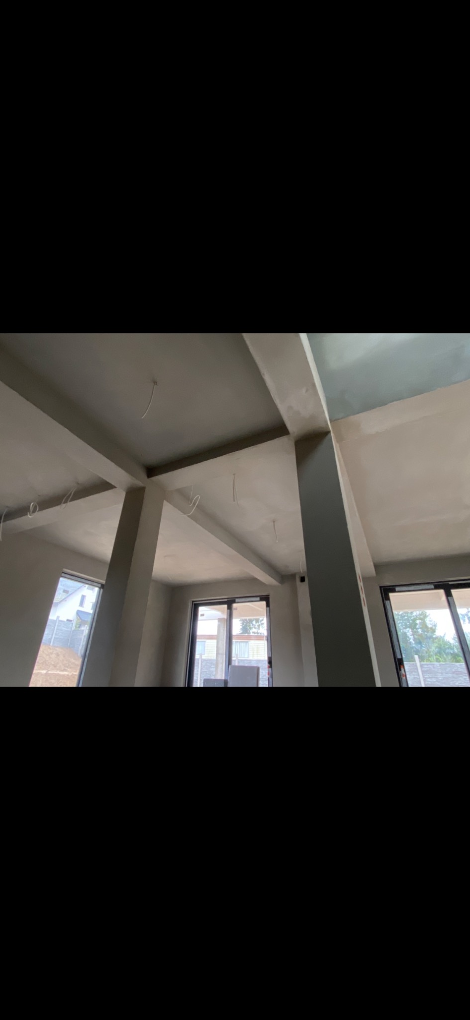 Surowe wnętrze budynku w trakcie prac wykończeniowych: widoczne szare ściany, betonowe filary, okna oraz wystające przewody elektryczne z sufitu.