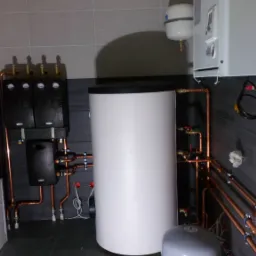 Kotłownia z białym zasobnikiem ciepłej wody, czarnymi rozdzielaczami i miedzianą instalacją, widoczne elementy kotła wiszącego.