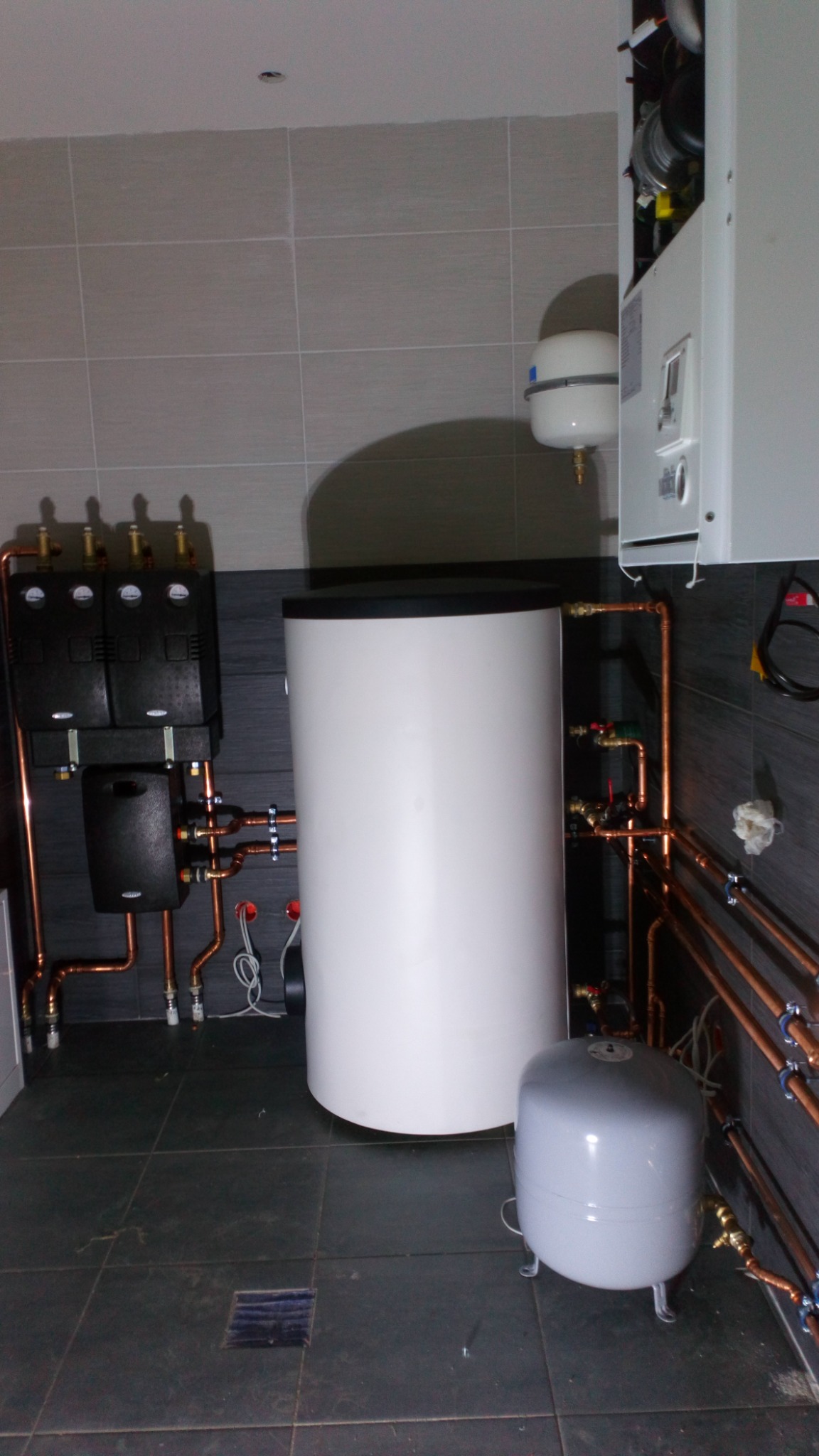 Kotłownia z białym zasobnikiem ciepłej wody, czarnymi rozdzielaczami i miedzianą instalacją, widoczne elementy kotła wiszącego.