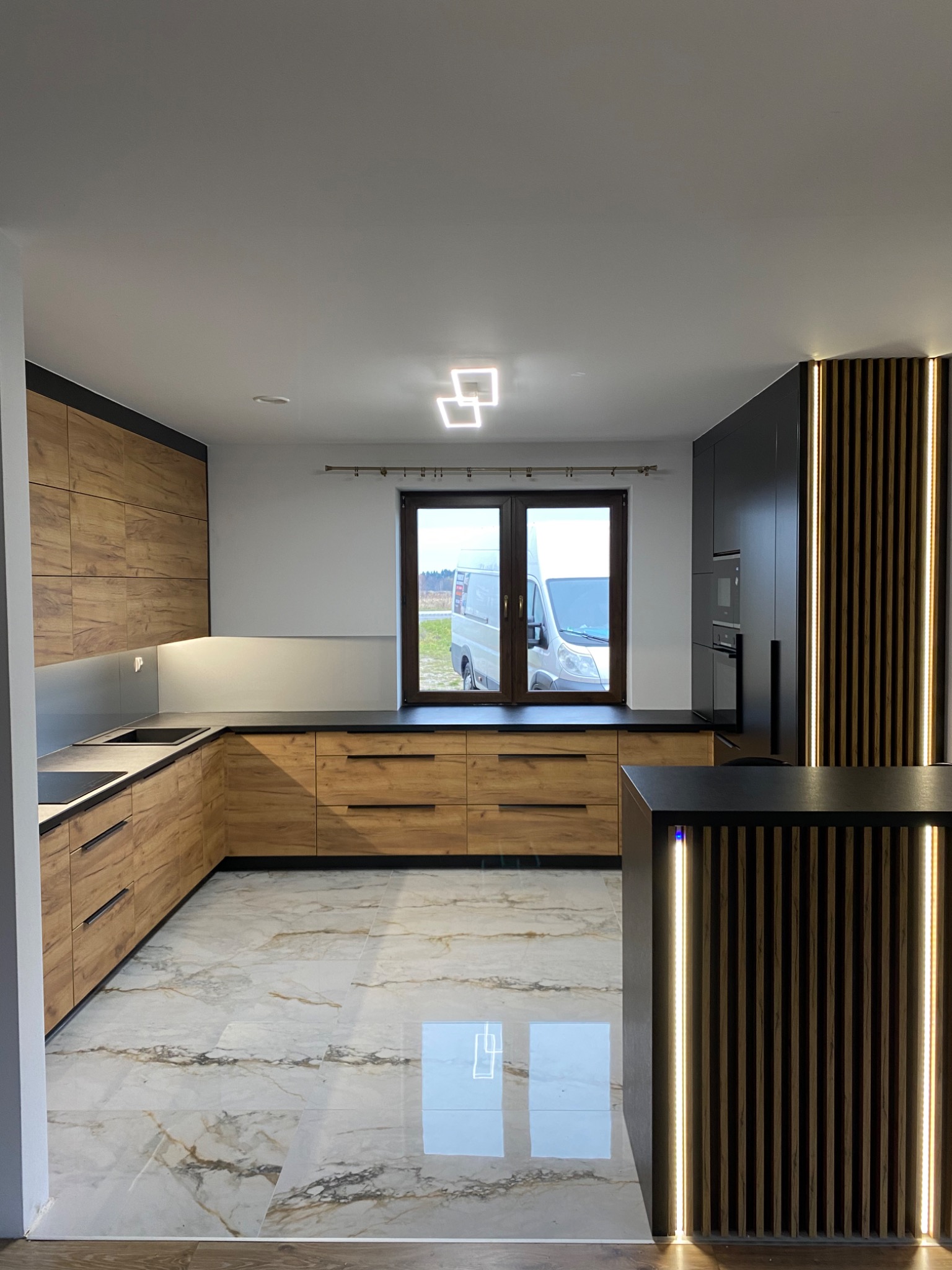 Nowoczesna kuchnia z drewnianymi frontami szafek, czarnym blatem i podświetlanymi lamelami na wyspie, widok na samochód dostawczy za oknem.
