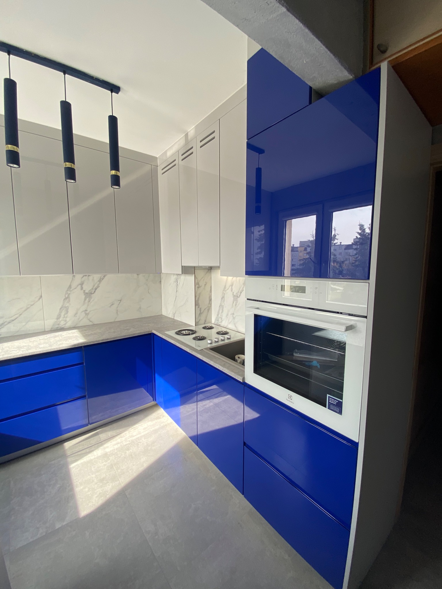 Nowoczesna kuchnia z błyszczącymi, granatowymi frontami szafek dolnych i białymi szafkami górnymi, marmurowy backsplash, oświetlenie punktowe i wbudowany piekarnik Electrolux.