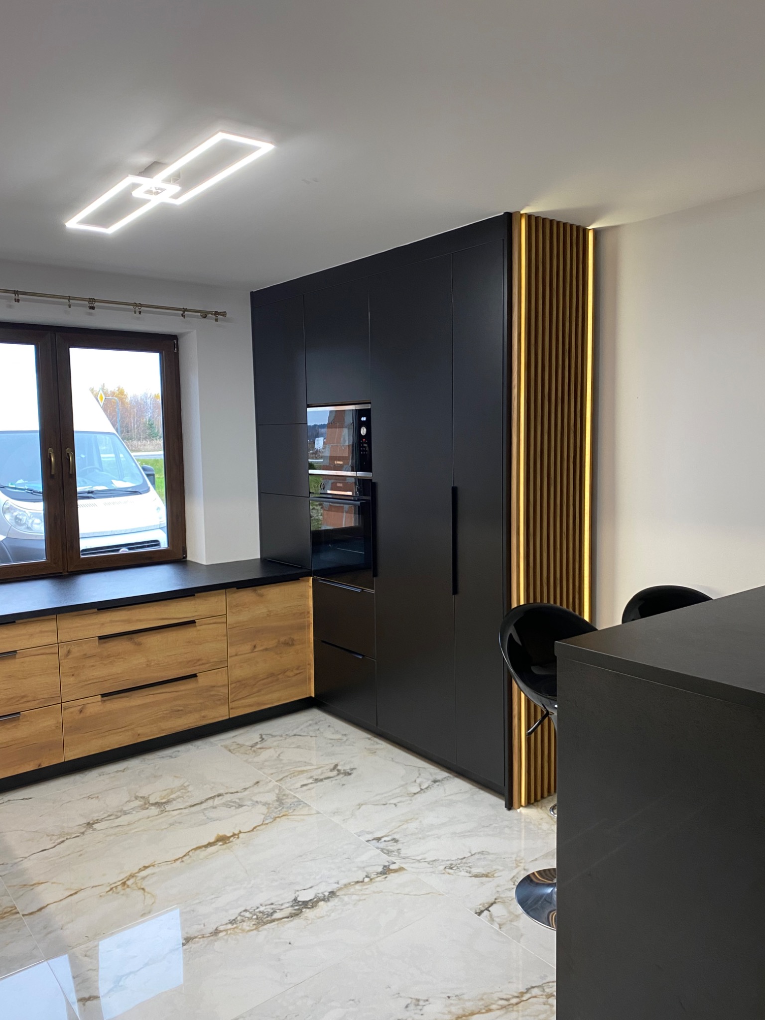 Nowoczesna kuchnia: czarne szafki z piekarnikiem, drewniane szuflady pod oknem, dekoracyjny panel z drewnianych listew z podświetleniem LED i marmurowe płytki podłogowe.