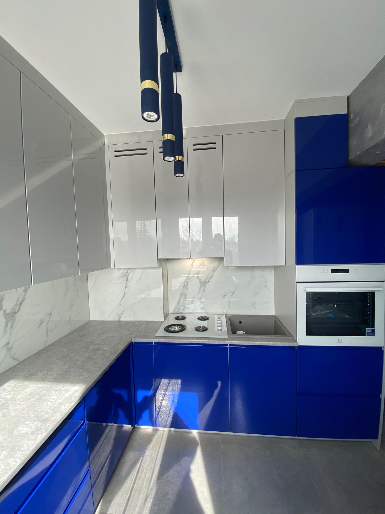 Nowoczesna kuchnia z białymi i niebieskimi lakierowanymi frontami, marmurowym blatem i backsplashiem, oświetlona trzema podłużnymi lampami w kolorze granatowym ze złotymi akcentami.