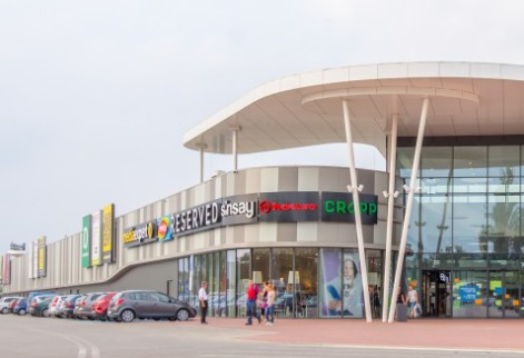 Nowoczesna hala handlowa z widocznymi szyldami sklepów: Media Expert, Reserved, Sinsay, House, Cropp, z zaparkowanymi samochodami i przechodzącymi ludźmi w słoneczny dzień.