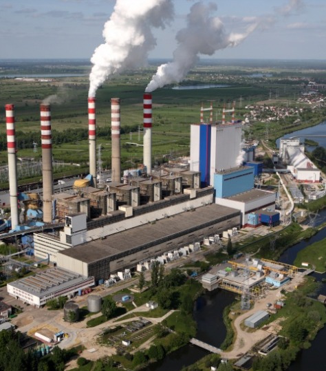Panorama elektrowni z kominami w biało-czerwone pasy, z dymem unoszącym się w górę, otoczonej zielenią i rzeką, widok z lotu ptaka.