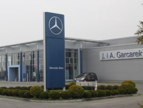 Nowoczesny budynek salonu samochodowego Mercedes-Benz z dużym pylonem reklamowym, otoczony ozdobnymi krzewami.