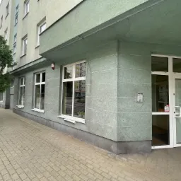 Zewnętrzny widok biurowca z wejściem, zielona elewacja z poziomymi pasami, białe okna i drzwi, kostka brukowa na chodniku, drzewo liściaste po lewej stronie.