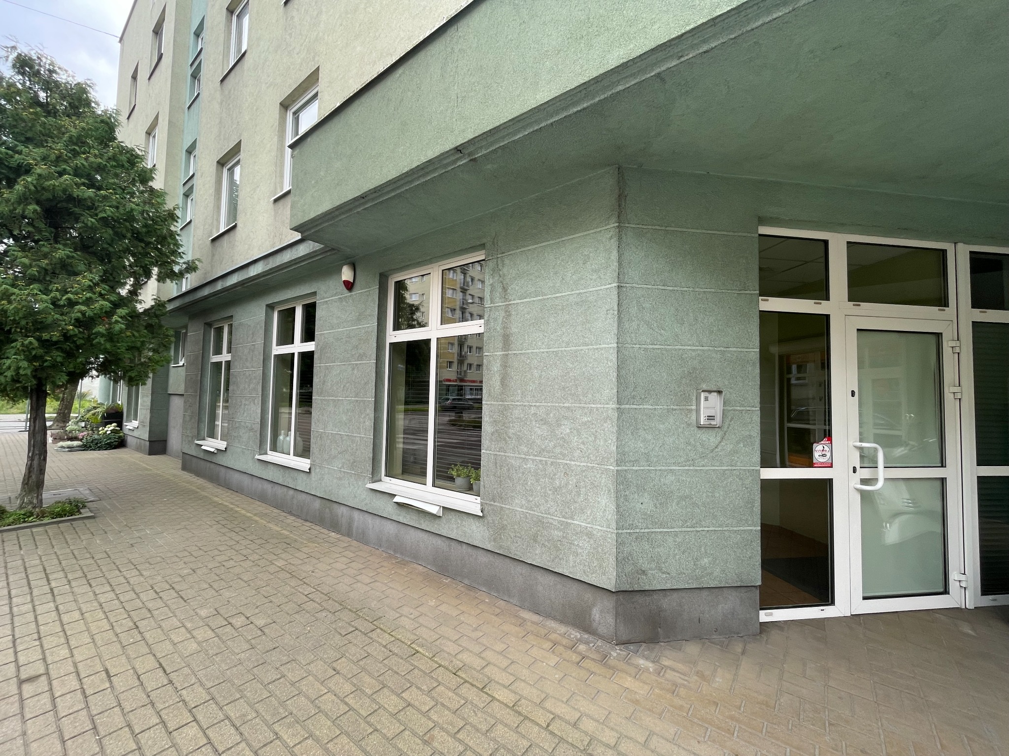 Zewnętrzny widok biurowca z wejściem, zielona elewacja z poziomymi pasami, białe okna i drzwi, kostka brukowa na chodniku, drzewo liściaste po lewej stronie.