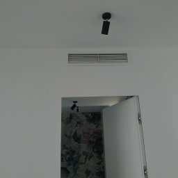 Klimaver - Biała ściana z zamontowaną kratką wentylacyjną i reflektorem sufitowym, widok na otwarte drzwi do pomieszczenia z tapetą w kwiaty.