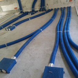 Klimaver - Niebieskie, karbowane rury wentylacyjne ułożone na surowym, betonowym podłożu w trakcie instalacji rekuperacji w budynku. Rury przymocowane metalowymi obejmami, prowadzą do niebieskich skrzynek...