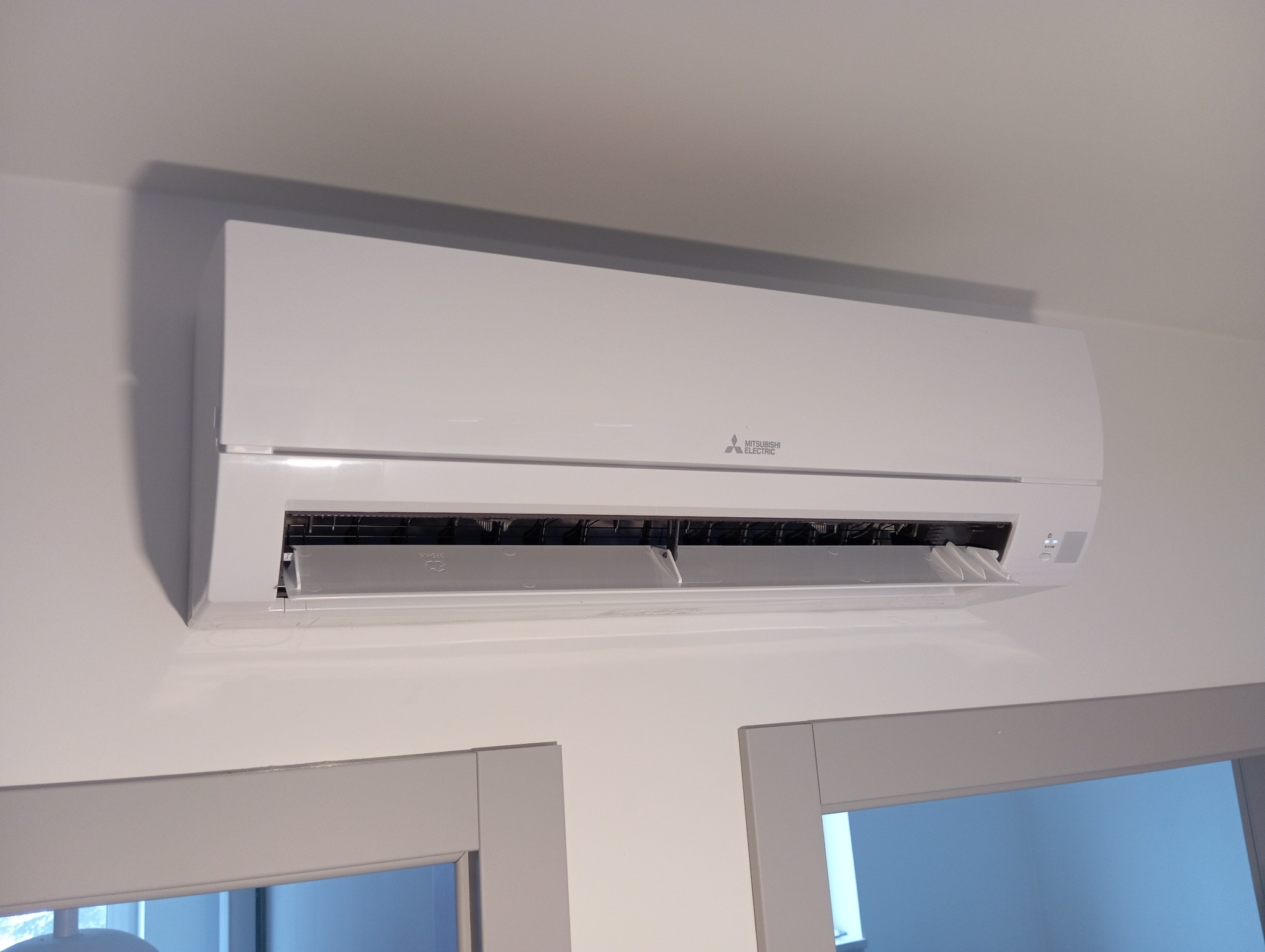 Biała jednostka wewnętrzna klimatyzacji Mitsubishi Electric zamontowana na ścianie nad szarą framugą drzwi, widoczne otwarte żaluzje wylotu powietrza.