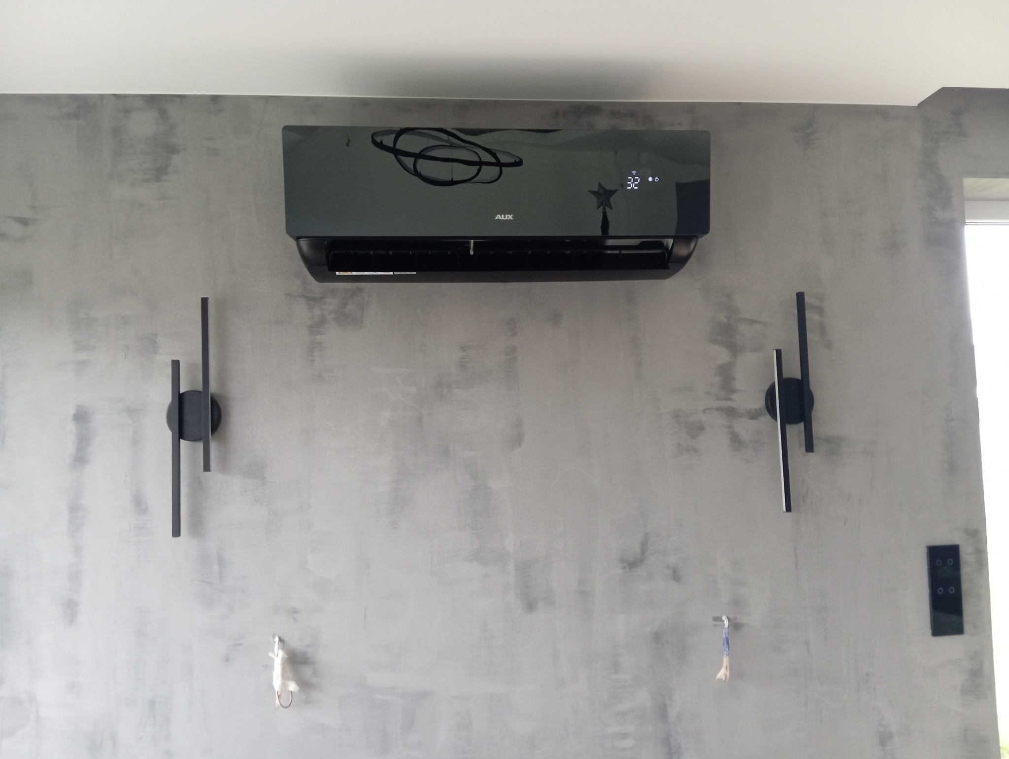 Czarny klimatyzator AUX zamontowany na szarej ścianie o fakturze betonu, z widocznym wyświetlaczem temperatury 32 stopnie, obok dwie nowoczesne lampy ścienne w stylu minimalistycznym.