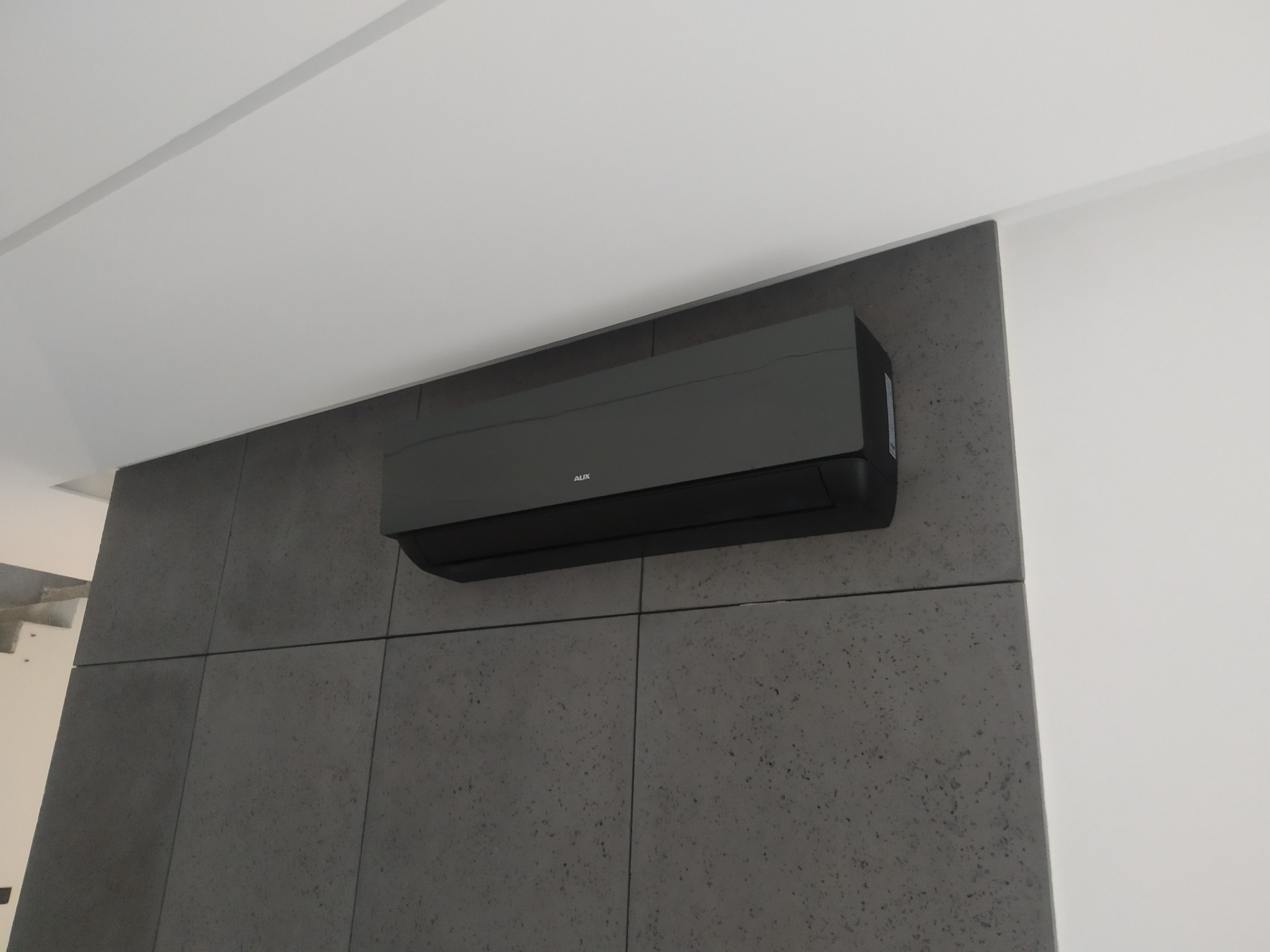 Czarny klimatyzator marki AUX zamontowany na szarej, teksturowanej ścianie w mieszkaniu w Warszawie, widok z dołu.