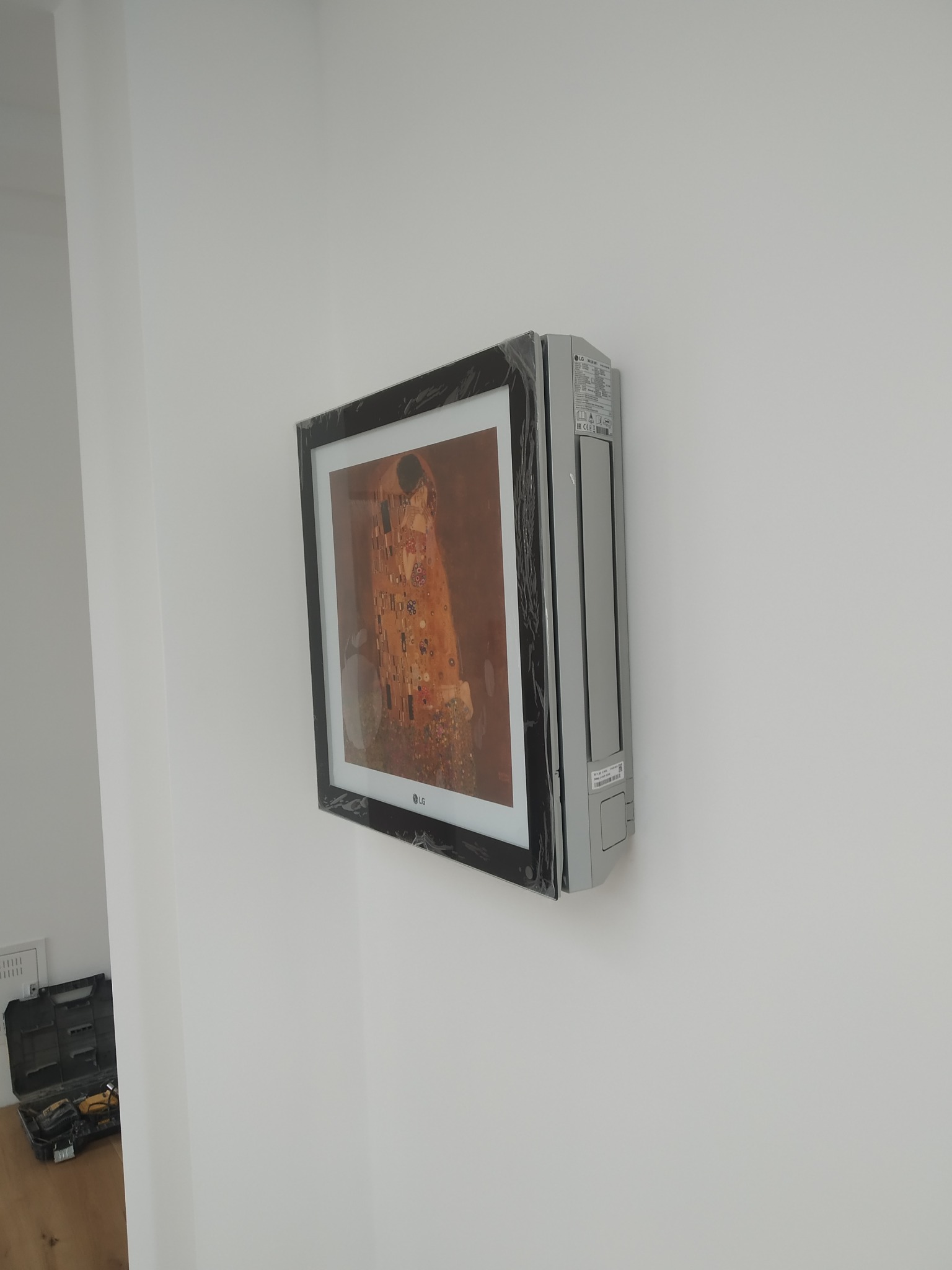 Klimatyzator LG Artcool Gallery w folii ochronnej, z reprodukcją obrazu 'Pocałunek' Gustava Klimta, zamontowany na białej ścianie, widoczny w tle czarny kufer z narzędziami.