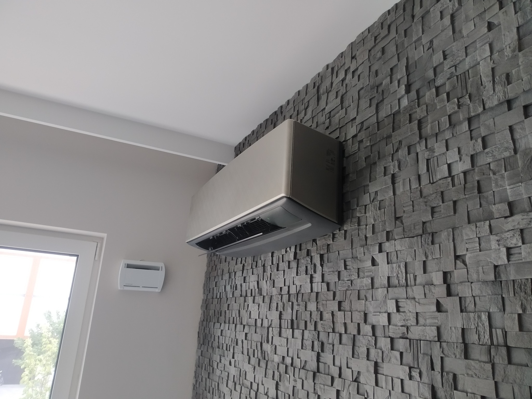 Szary klimatyzator ścienny zamontowany na dekoracyjnej ścianie z kamiennych kostek, widoczny z dołu, z oknem i panelem sterowania w tle.