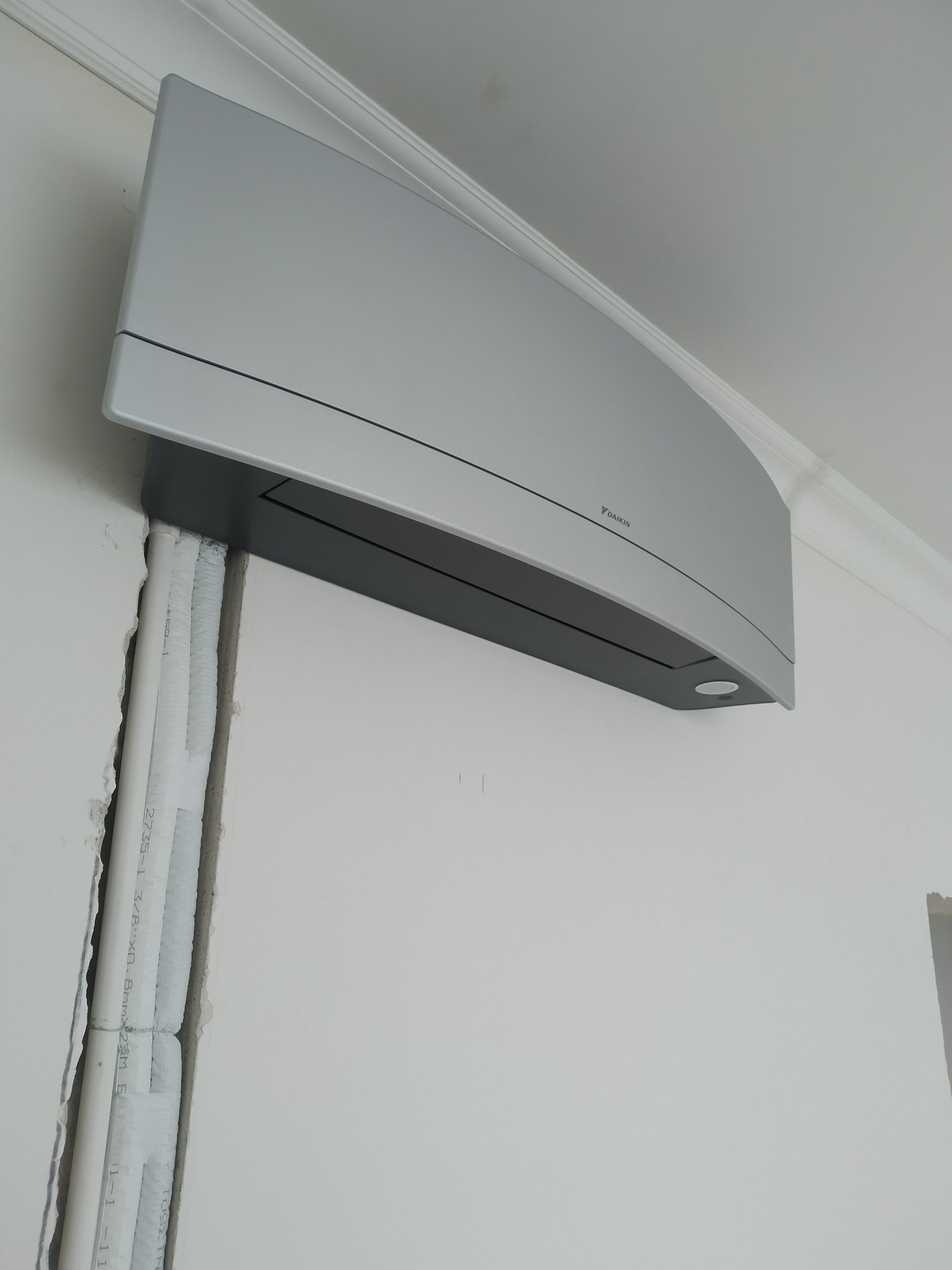 Szary klimatyzator Daikin zamontowany na białej ścianie, widoczne rury instalacyjne w bruździe ściennej.