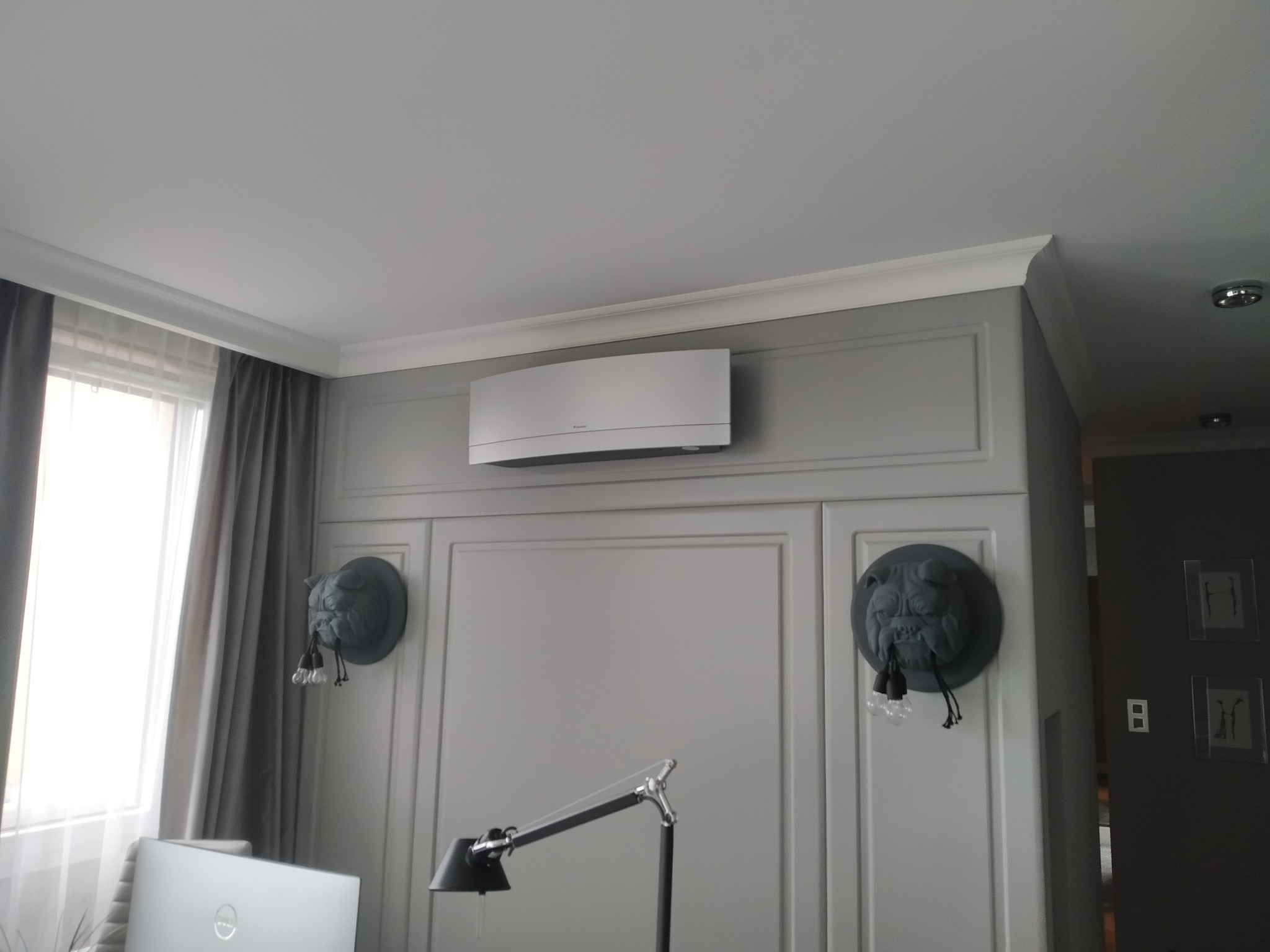 Nowoczesny klimatyzator ścienny Daikin w szarej obudowie, zamontowany nad ozdobną ścianą z panelami i dekoracyjnymi lampami w kształcie buldogów, oświetlony lampą biurkową.