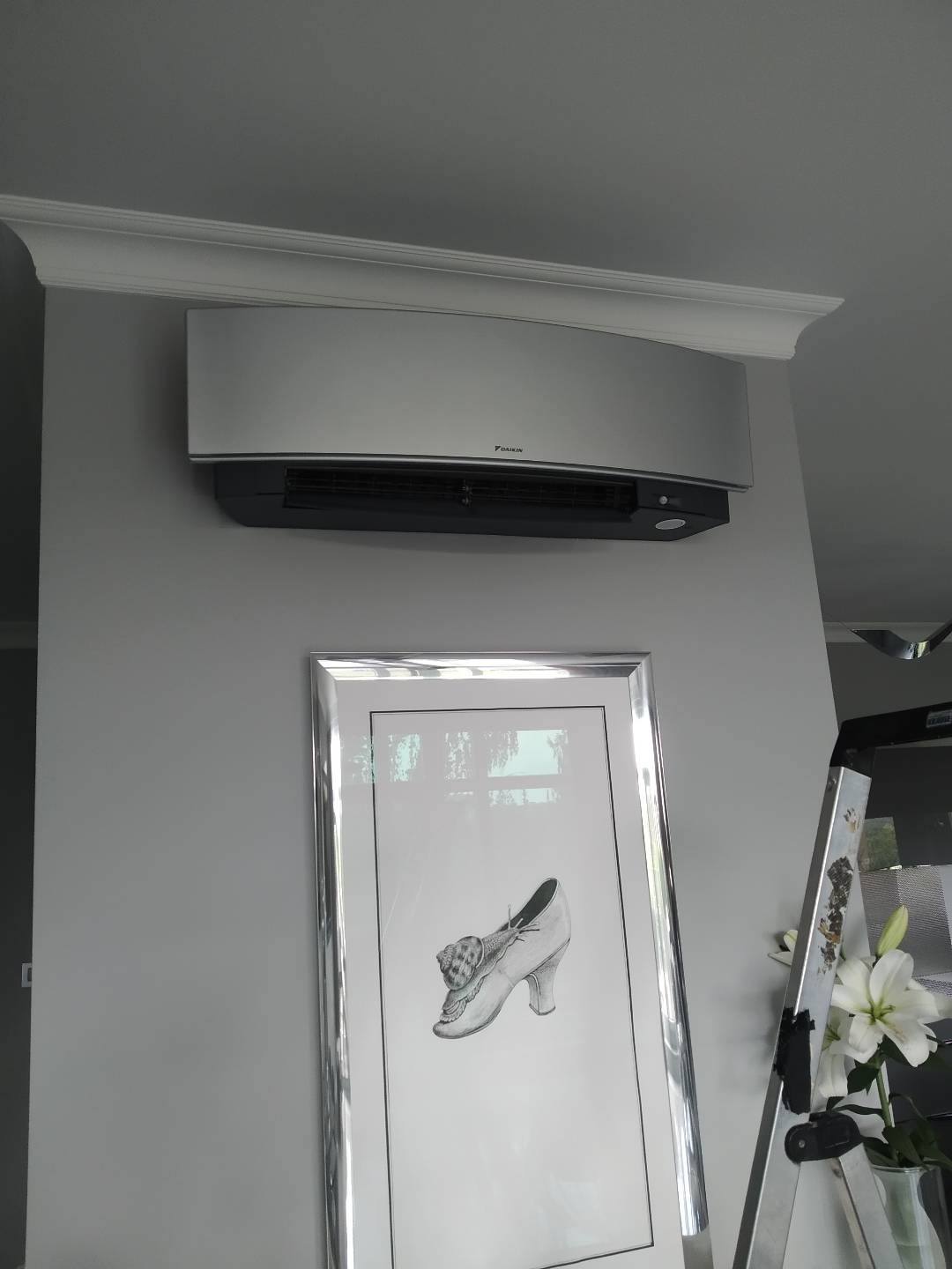 Szary klimatyzator Daikin zamontowany pod sufitem z dekoracyjną listwą, pod nim obraz w srebrnej ramie przedstawiający buta ze ślimakiem, fragment drabiny i wazon z liliami po prawej stronie.