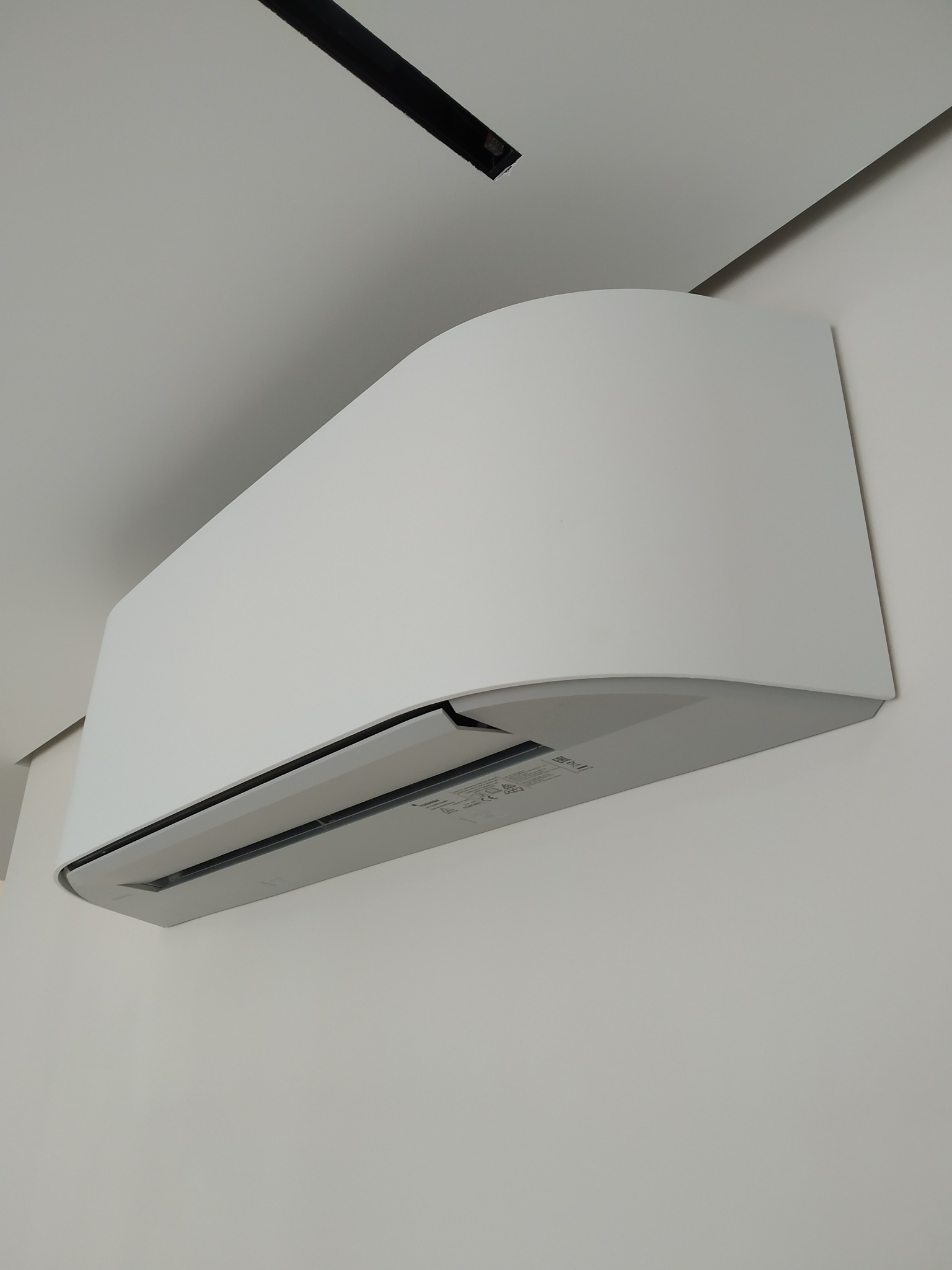 Biała jednostka wewnętrzna klimatyzacji, zamontowana na ścianie, widoczna z perspektywy dolnej, z fragmentem sufitu i elementami instalacji elektrycznej w tle.