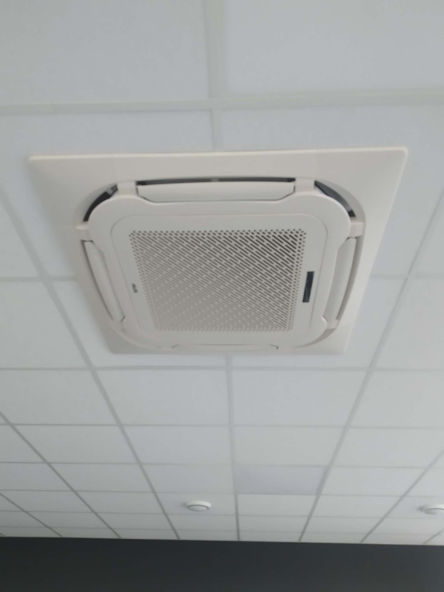 Biała, sufitowa kaseta klimatyzacyjna z kratką wentylacyjną, zamontowana w biurze z podwieszanym sufitem kasetonowym.