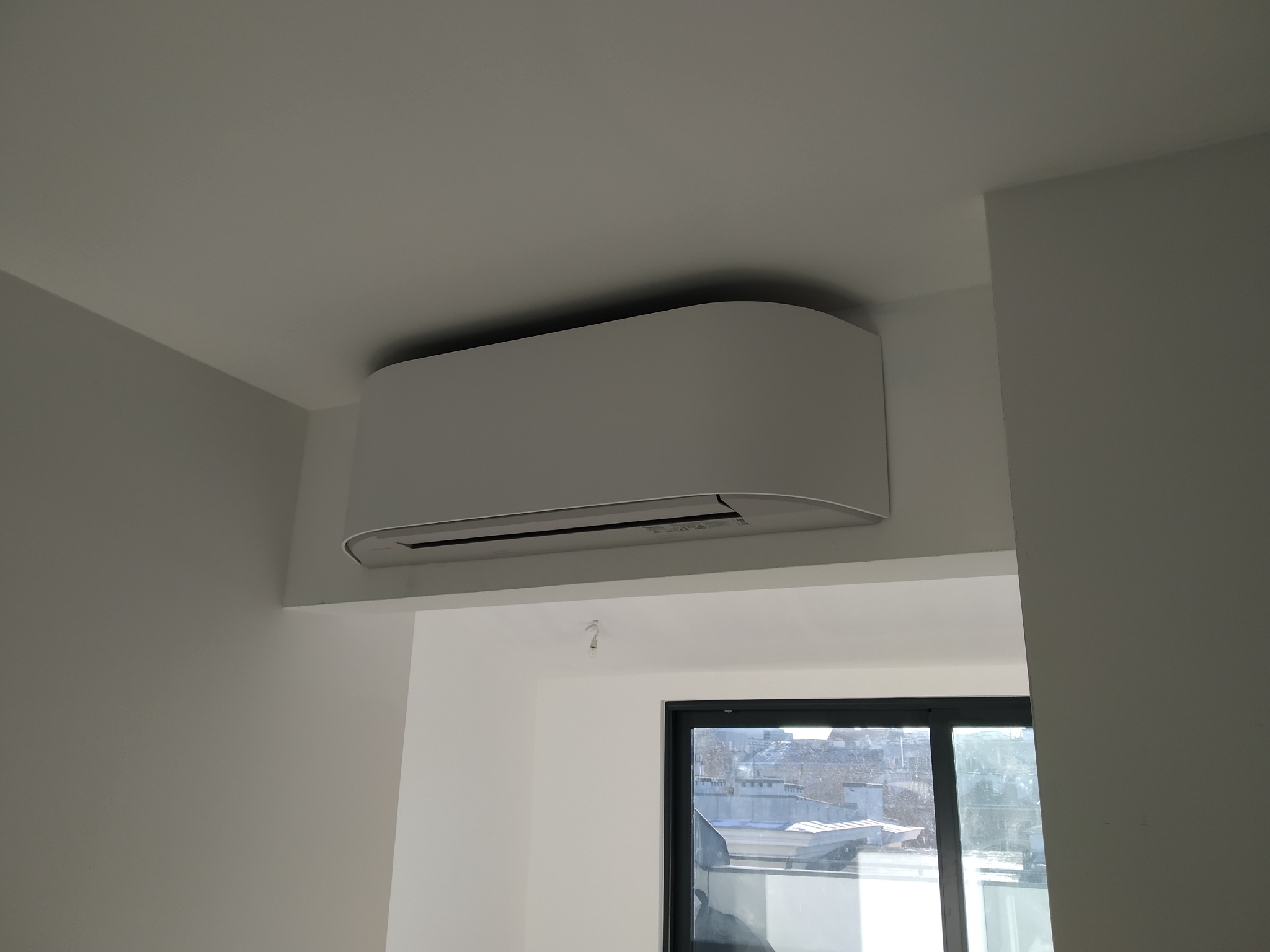 Biała klimatyzacja ścienna, zamontowana w rogu pokoju z widokiem na miasto przez okno.