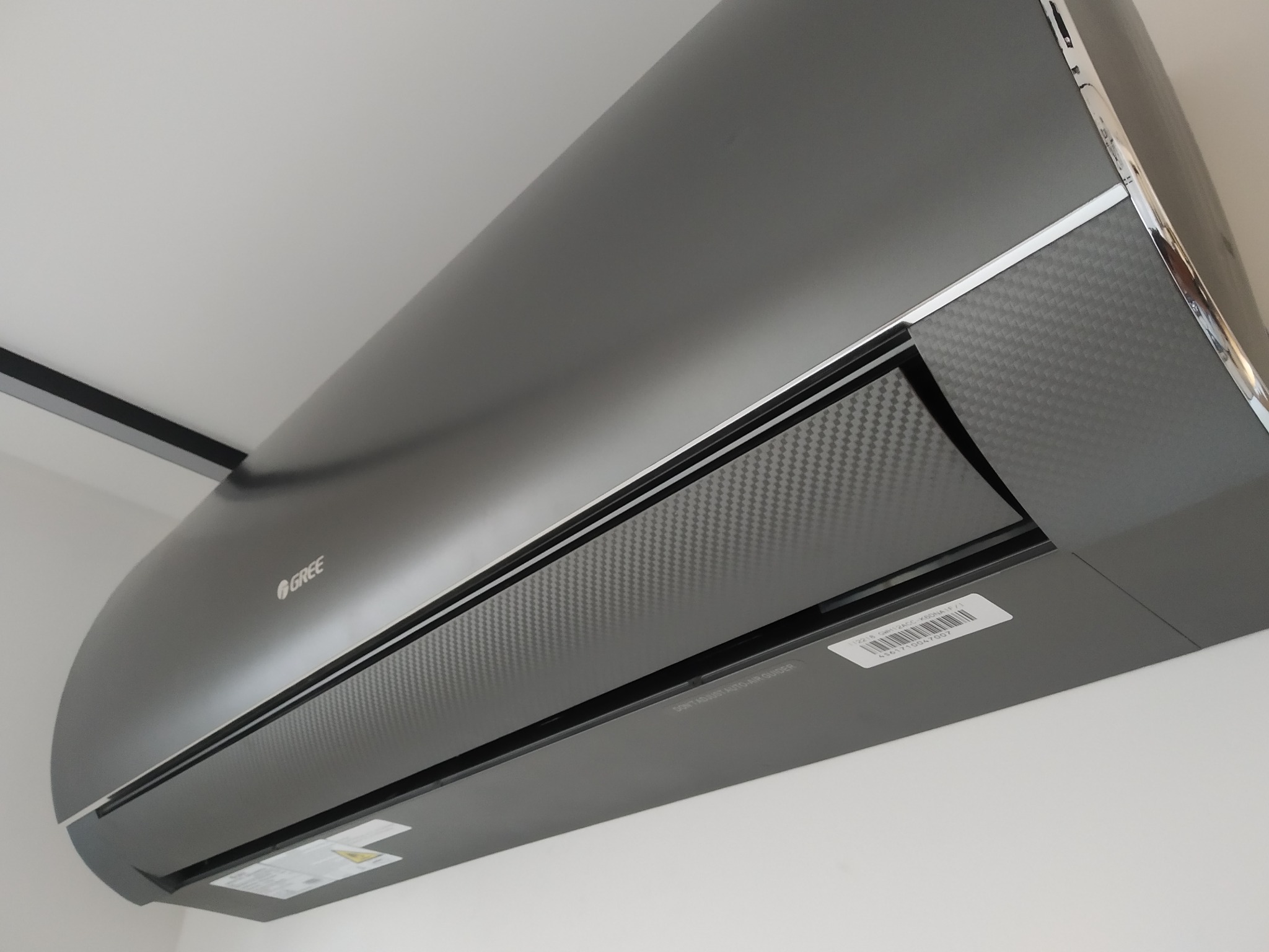 Czarny klimatyzator GREE z panelem o fakturze włókna węglowego zamontowany na białej ścianie, widoczny fragment czarnej listwy sufitowej.